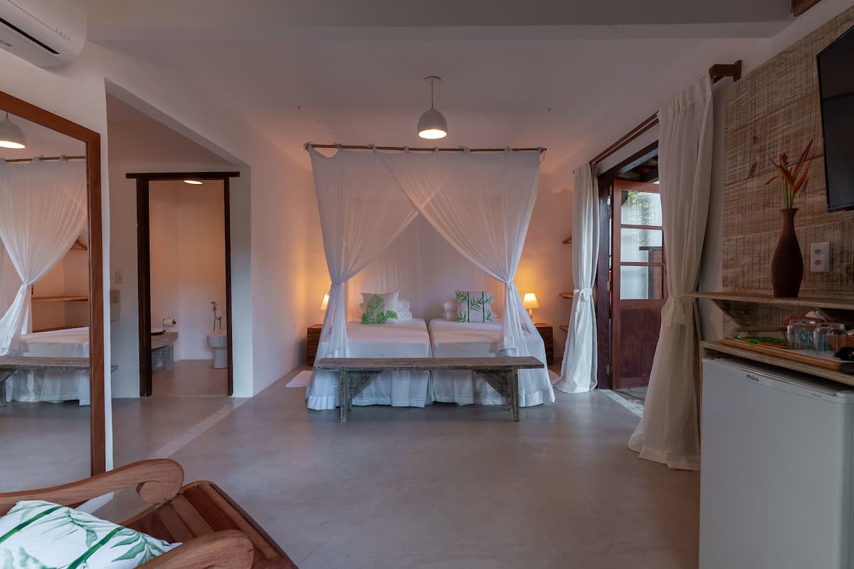 Un dormitorio con cama con dosel, un banco, un refrigerador y una mesita de noche con un florero.