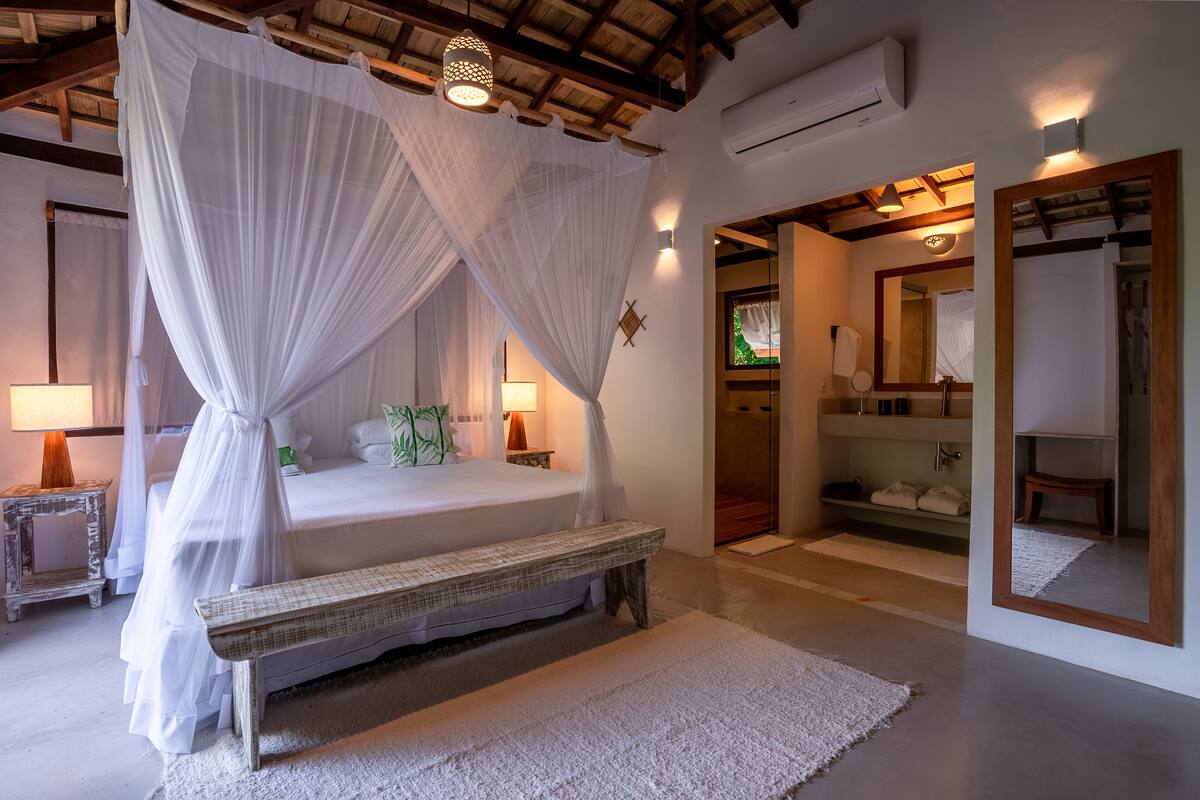 Un dormitorio con cama con dosel, lámparas de noche, un banco, un espejo y un vestidor.