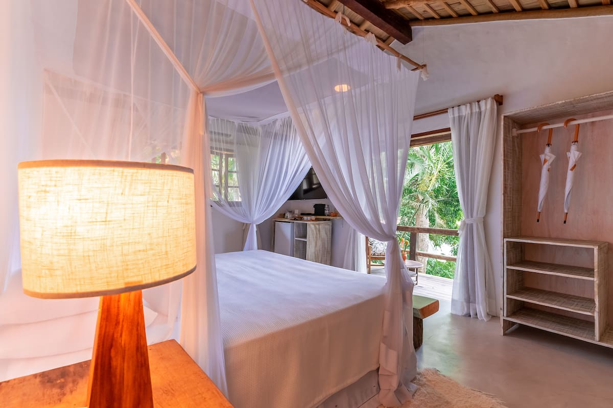 Un dormitorio con cama a dosel, lámpara de noche, mesita de madera y vistas a la vegetación exterior.