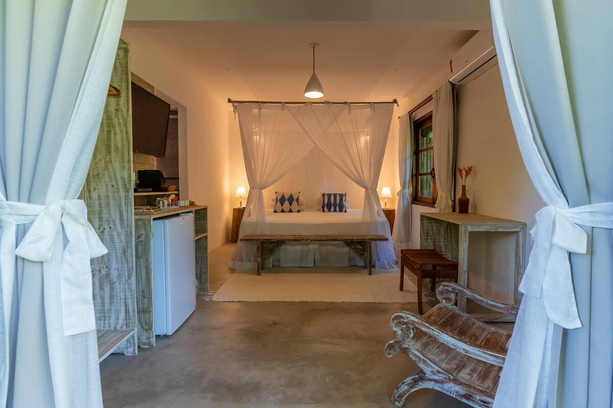 Un dormitorio con cama, lámparas de noche, mesitas de noche, un banco y televisión sobre un mueble.