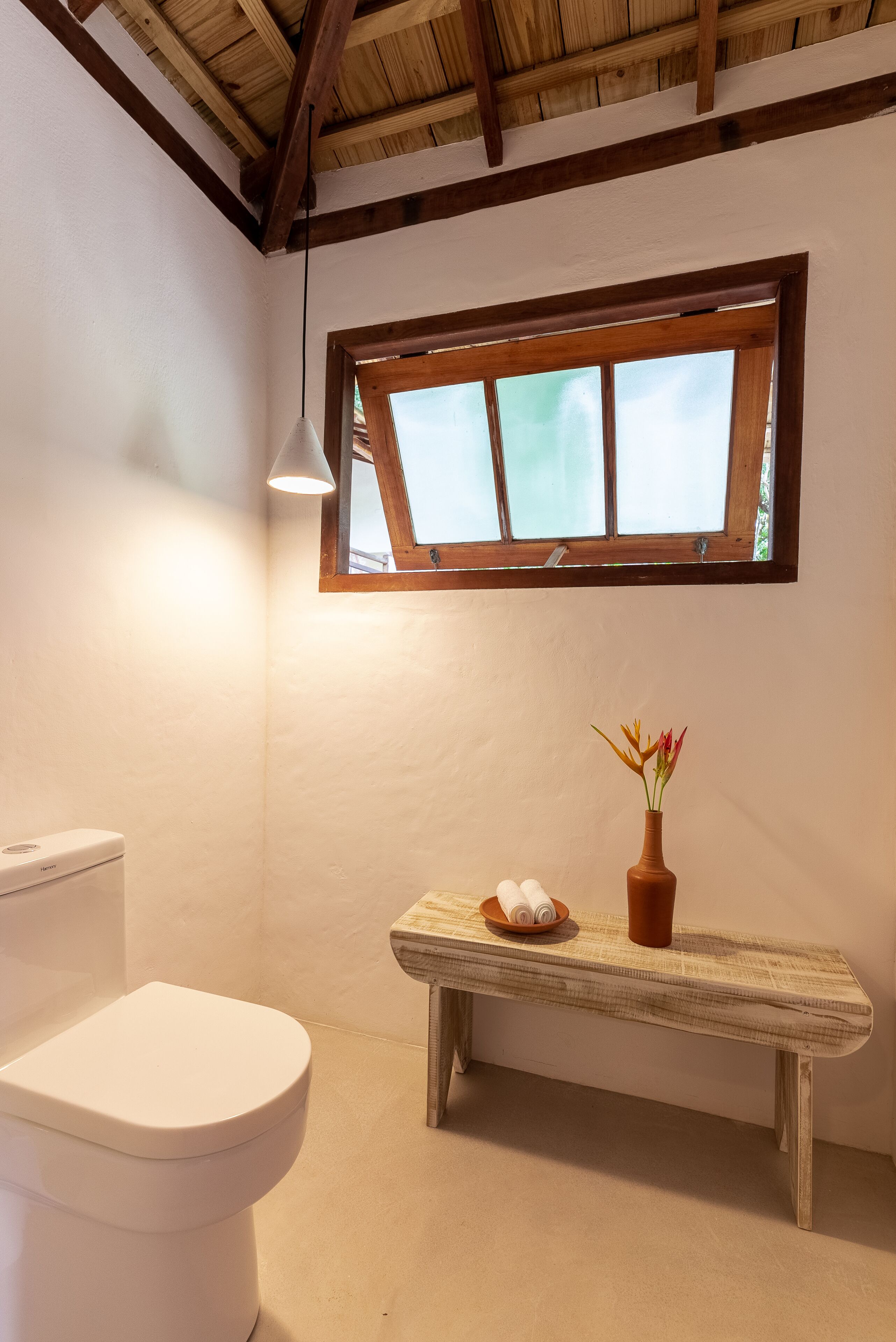 A bathroom with a white toilet, a wooden bench with a vase of flowers, and a window with a wooden frame.