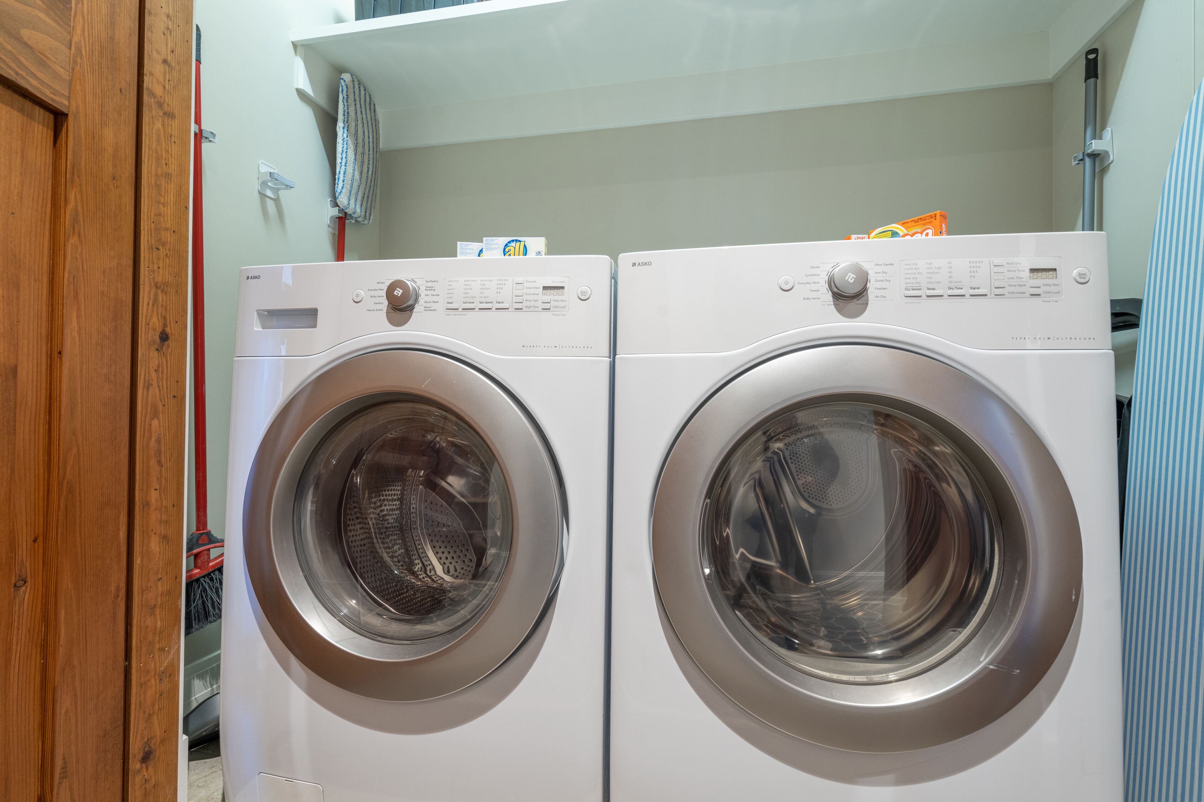 A pair of front-loading washing machines in a laundry room with a wooden door and a mop on the floor.