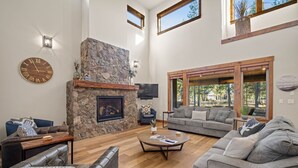 A spacious living room with a stone fireplace, comfortable seating, and large windows offering a view of the outdoors. - Family Getaway with Access to Forest House (Sunriver)