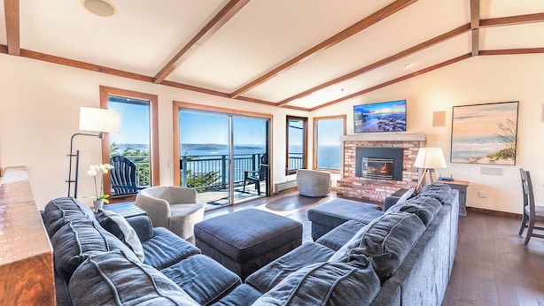 Een ruime woonkamer met een grote grijze modulaire bank, een open haard en een balkon met uitzicht op de oceaan.