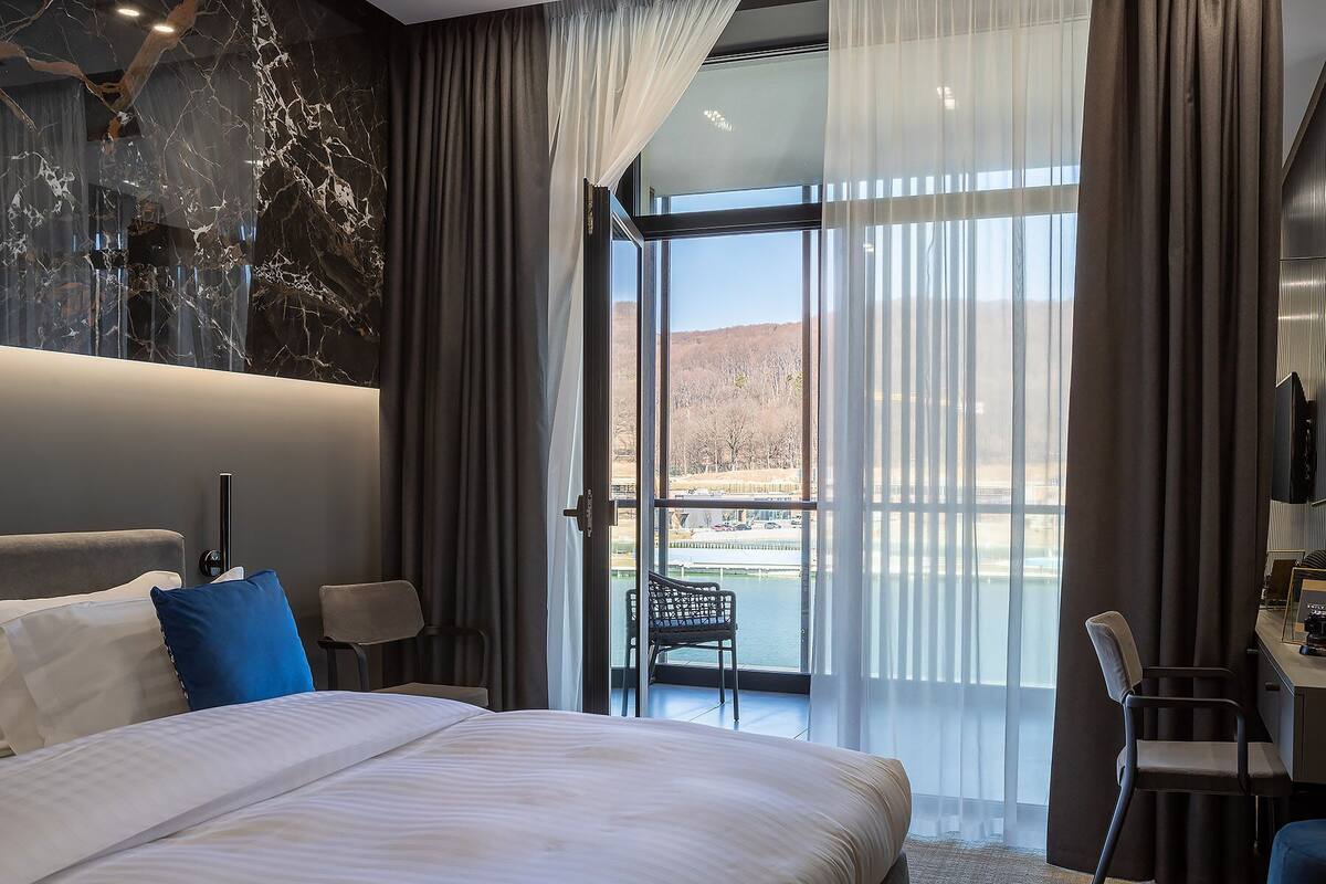 Quarto de hotel moderno com uma cama grande, uma escrivaninha e vista para a água e colinas.