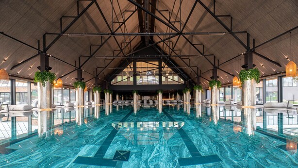 Una amplia piscina cubierta con techos altos y diseño moderno, rodeada de zonas de estar y grandes ventanales.