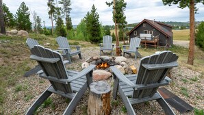 A fire pit with four Adirondack chairs arranged around it, a wooden deck, and a cabin in the background.