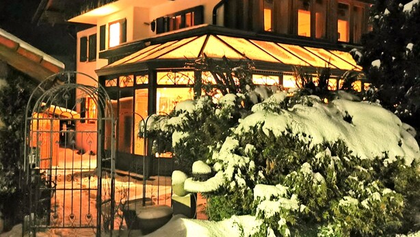 Una casa moderna de dos pisos con un amplio invernadero de vidrio, un paisaje cubierto de nieve y una entrada bien iluminada.