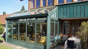 Un invernadero con techo de cristal, una zona de patio con mesa y sillas, y vistas del paisaje circundante.