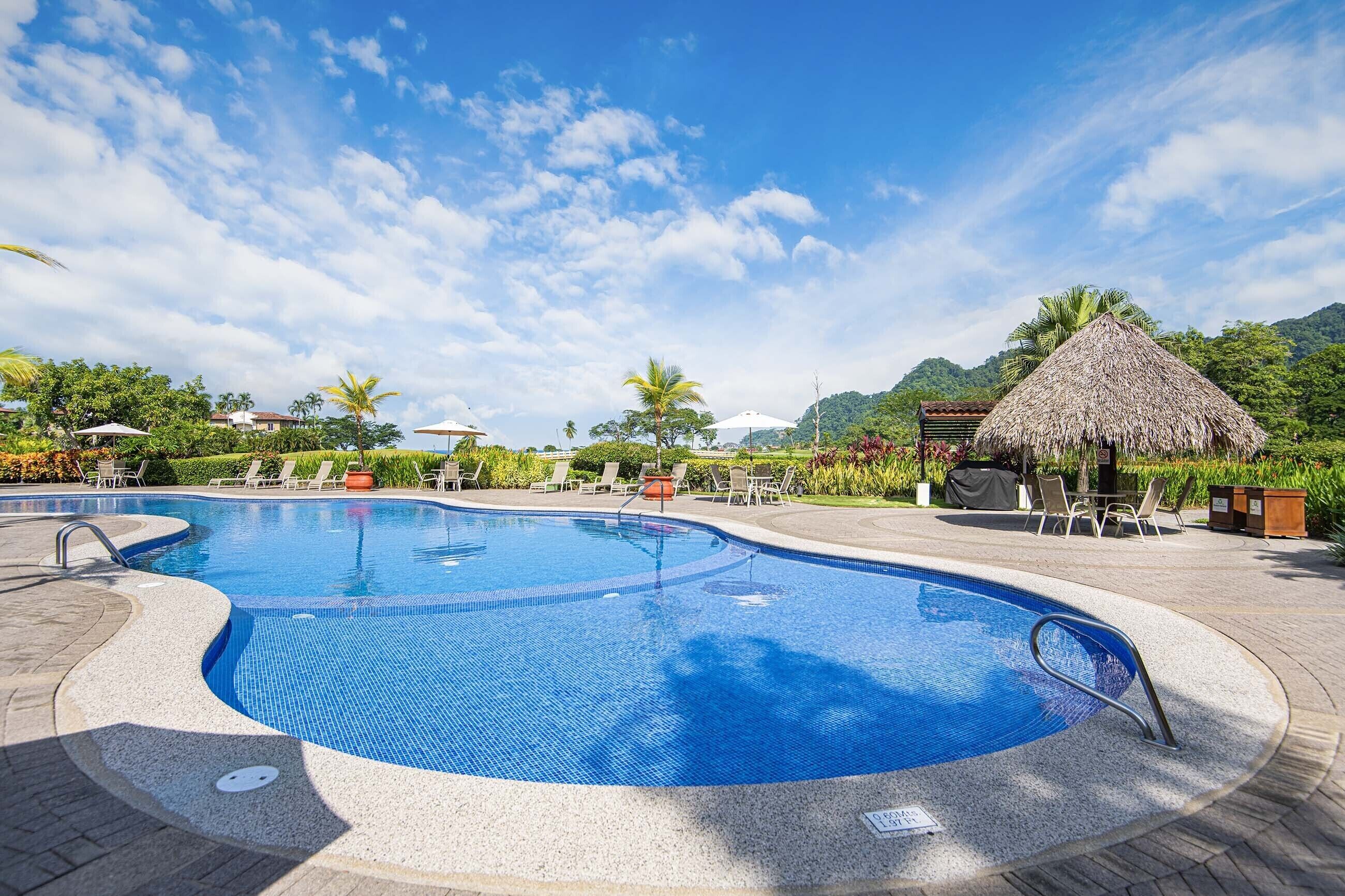 A swimming pool with a curved edge, surrounded by lounge chairs and umbrellas, set against a backdrop of lush greenery and a mountain.