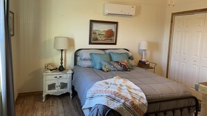A bedroom with a bed, bedside tables, a lamp, a painting, and an air conditioning unit.
