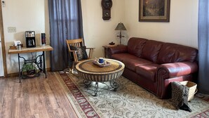 A living room with a leather sofa, a coffee table, a wooden chair, a small table with a lamp, a clock, and a painting on the wall.
