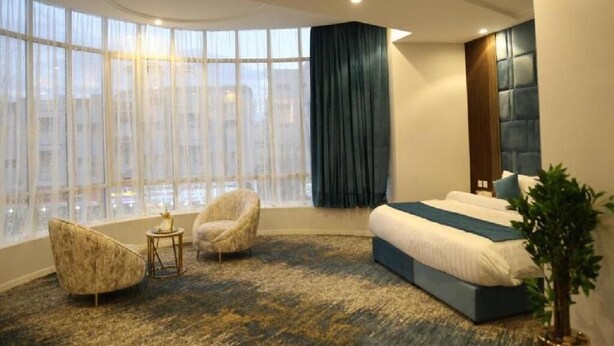 A modern hotel room with a large window, a curved ceiling, a bed with a blue headboard, and two armchairs. - Areen Hotel Airport (Jeddah)