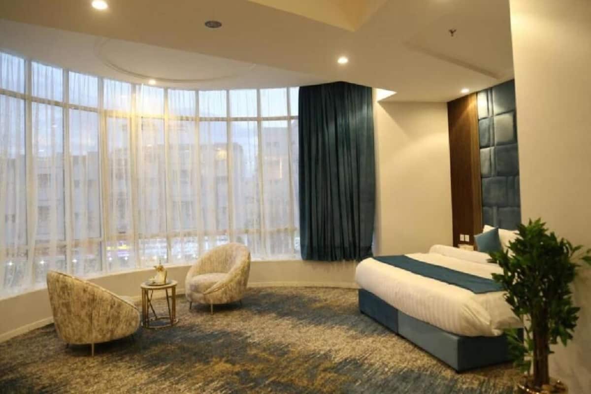 A modern hotel room with a large window, a curved ceiling, a bed with a blue headboard, and two armchairs.