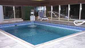 An indoor swimming pool with a tiled border, a lounge chair, and a view of city lights through large windows. - Areen Hotel Airport (Jeddah)