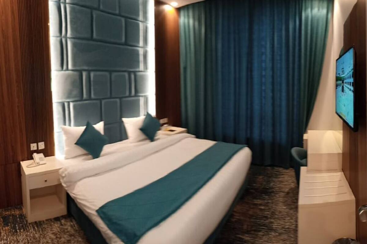 A hotel room with a large bed, a nightstand with a phone, a wall-mounted TV, and a blue curtain.