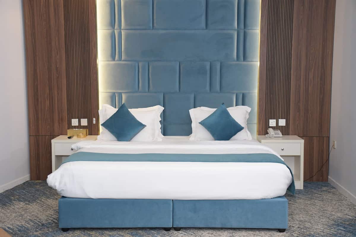 A modern hotel room with a large bed, a headboard with a geometric pattern, wooden paneling on the walls, and a small bedside table with a lamp and a phone.