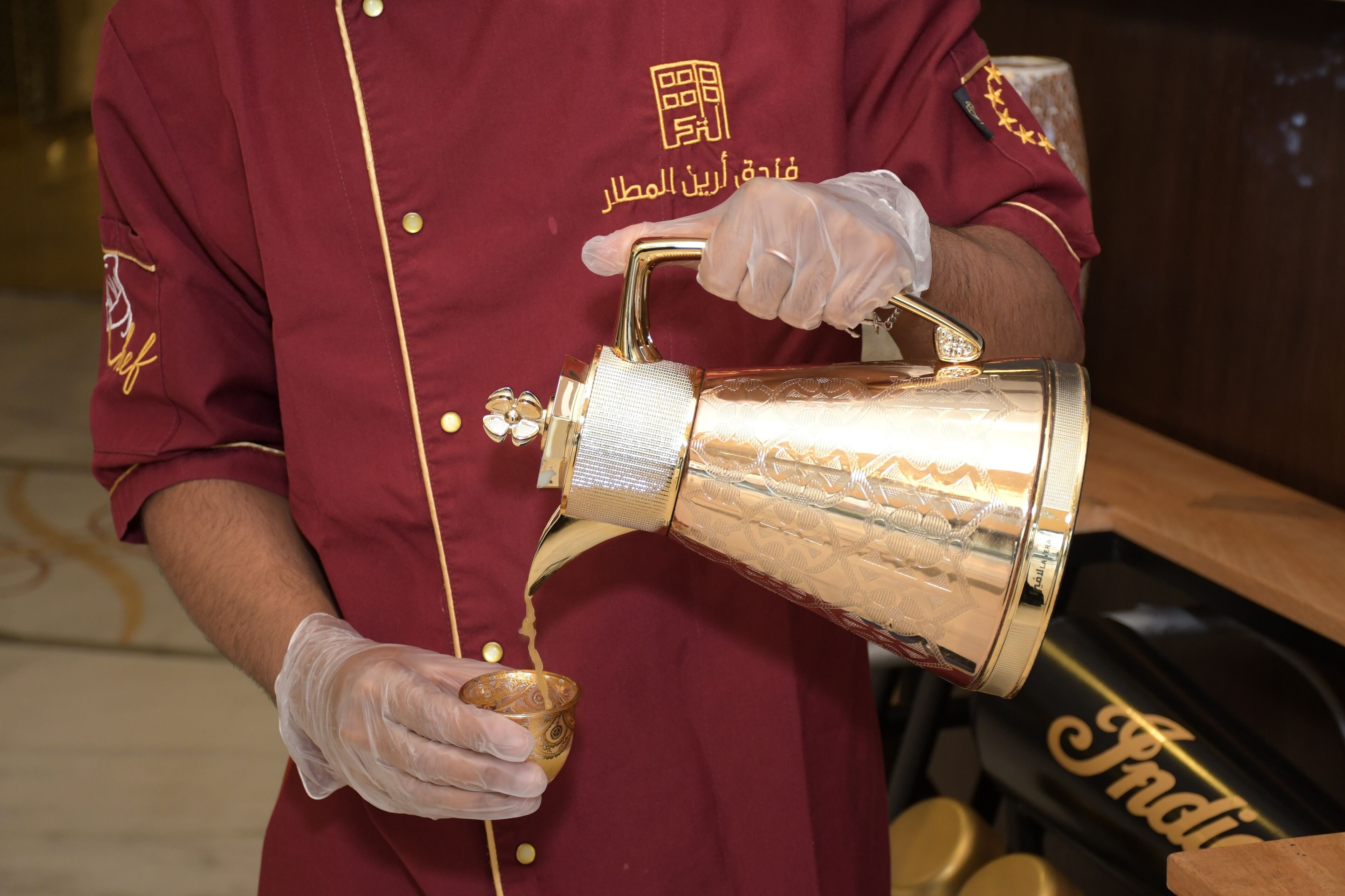 Una persona con uniforme color vino vertiendo un líquido dorado de un recipiente grande y ornamentado en una taza dorada más pequeña.