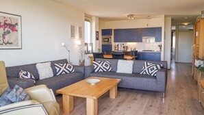 Een moderne woonkamer met een grijze bank, een houten salontafel en een keuken op de achtergrond.