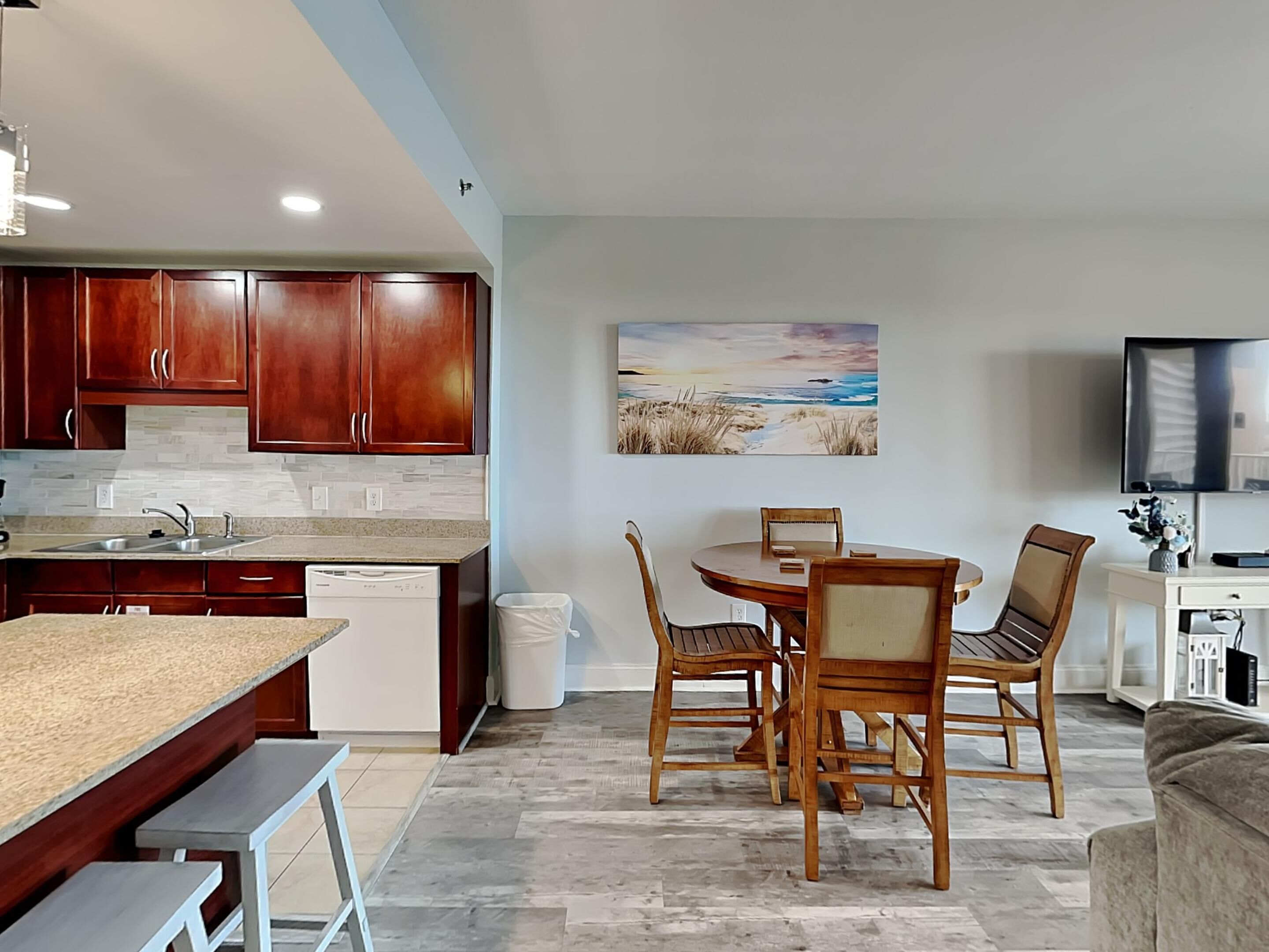 Una cocina moderna con armarios de madera, encimera de granito y lavavajillas integrado. Hay un comedor con mesa y sillas de madera, y un cuadro con una escena de playa en la pared.