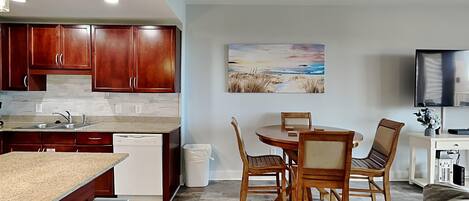 Una cocina moderna con armarios de madera, encimera de granito y lavavajillas integrado. Hay un comedor con mesa y sillas de madera, y un cuadro con una escena de playa en la pared.