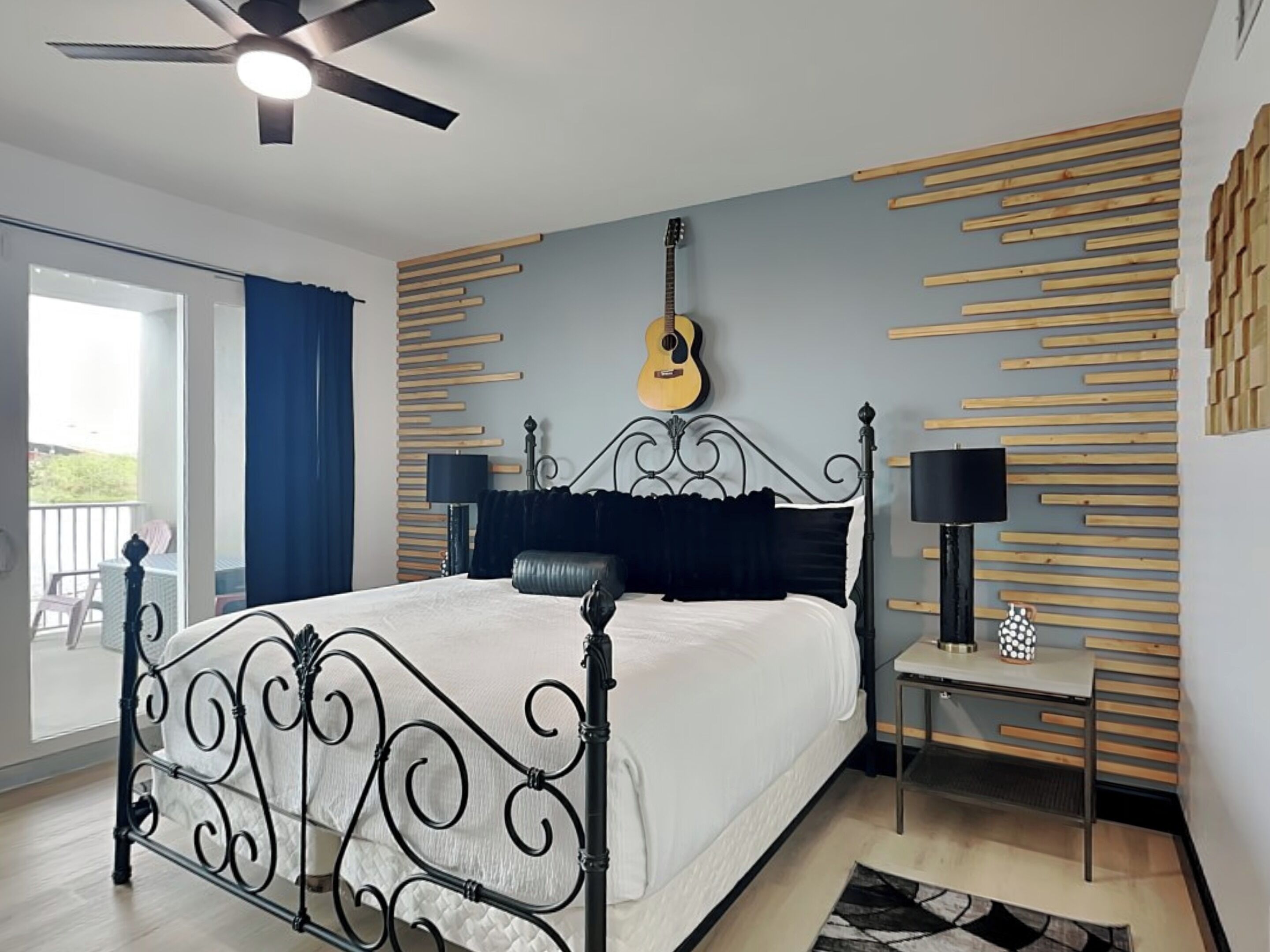 Quarto com cama, uma guitarra na parede, ventoinha no teto e varanda.