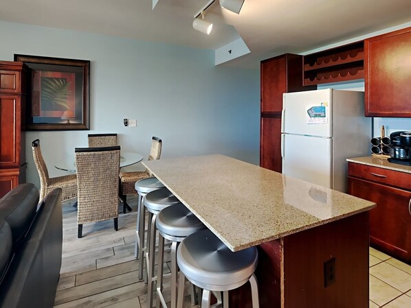 A kitchen with a granite countertop, bar stools, a refrigerator, and wooden cabinets. - Laketown Wharf by Resort Collection (Panama City Beach)