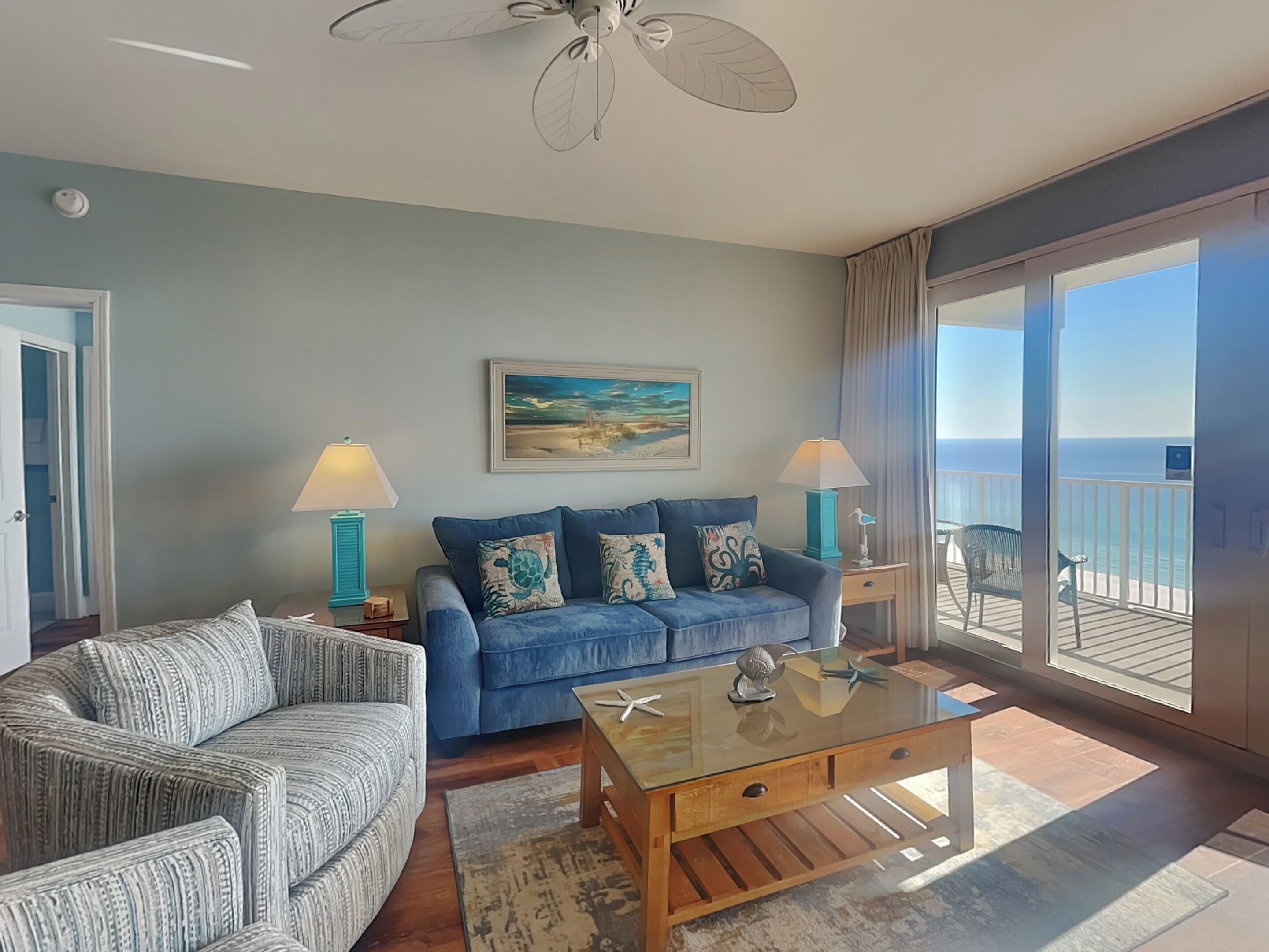 A living room with a blue sofa, a patterned armchair, a glass coffee table, and a painting of a beach scene.