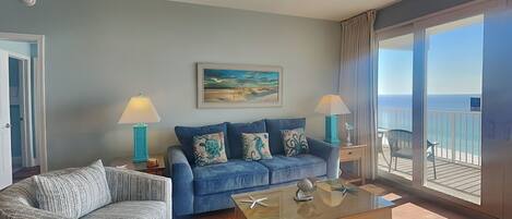 A living room with a blue sofa, a patterned armchair, a glass coffee table, and a painting of a beach scene.