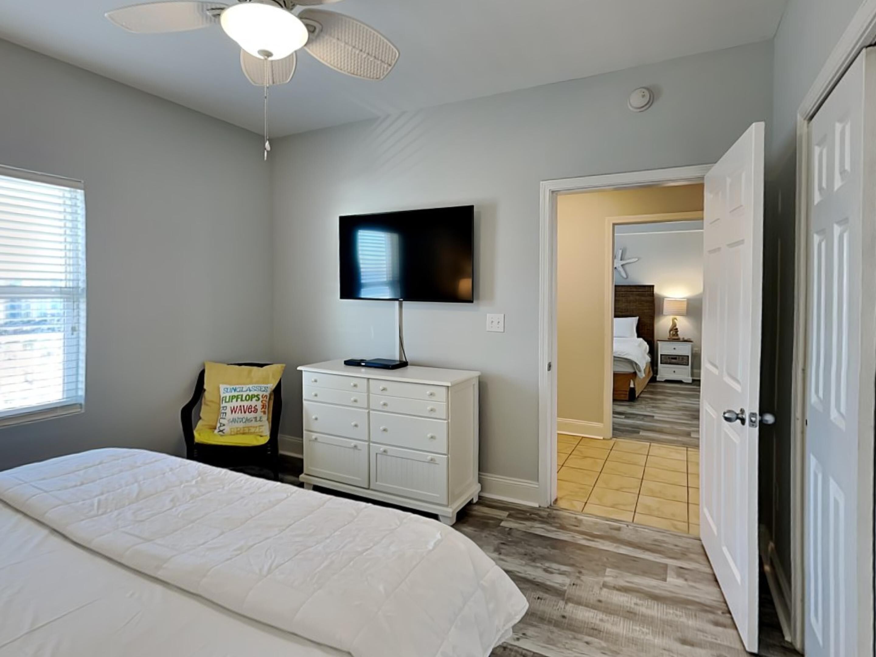 Un dormitorio con cama, cómoda blanca, ventilador de techo, televisor y puerta que da a otra habitación.