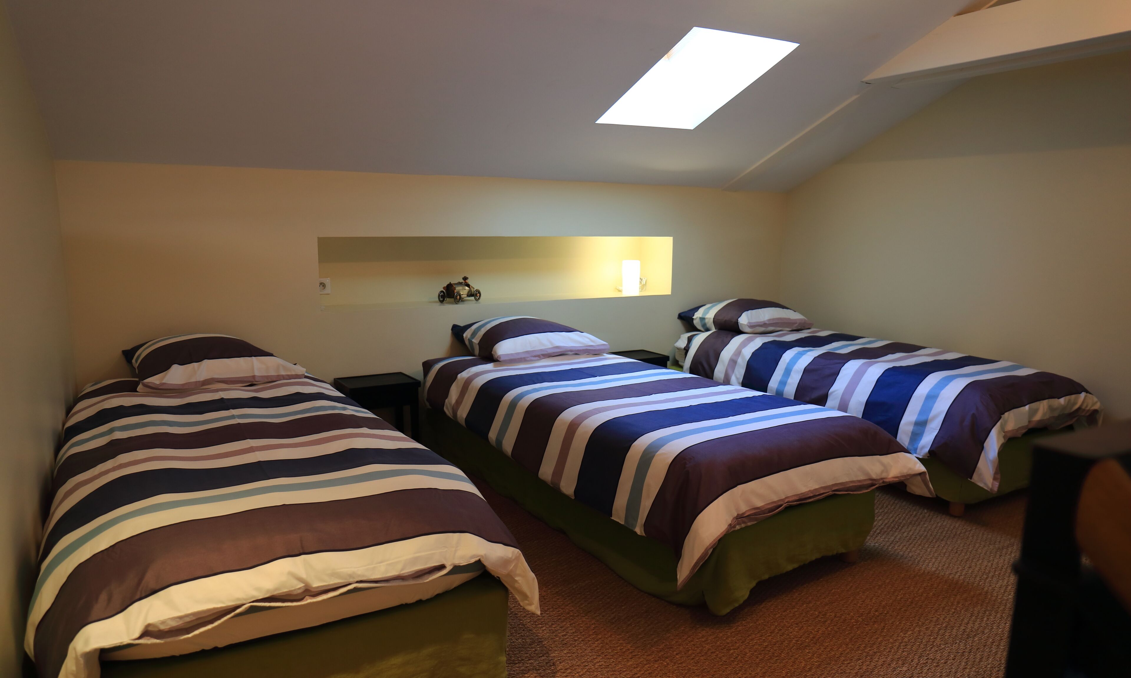 Una habitación con dos camas individuales, una mesita pequeña, un estante en la pared y un tragaluz.