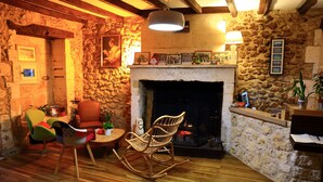 A cozy room with a stone wall, a fireplace, wooden flooring, and a variety of seating including a rocking chair and armchairs. - Hôtel Restaurant Le Jardin des chouchoux Brantôme  (Brantôme en Périgord)
