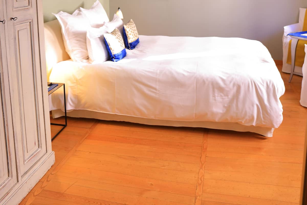 Una cama bien tendida con sábanas blancas y almohadones decorativos, un televisor montado en la pared y un piso de madera.