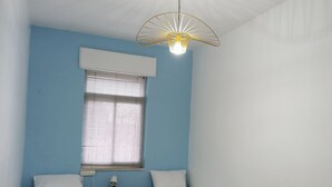 Une chambre avec deux lits simples, une fenêtre avec des stores, une petite table avec une lampe et un luminaire au plafond original.