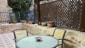 Une table ronde en verre, quatre chaises, un petit tabouret et plusieurs plantes en pot disposées sur un mur de pierre.