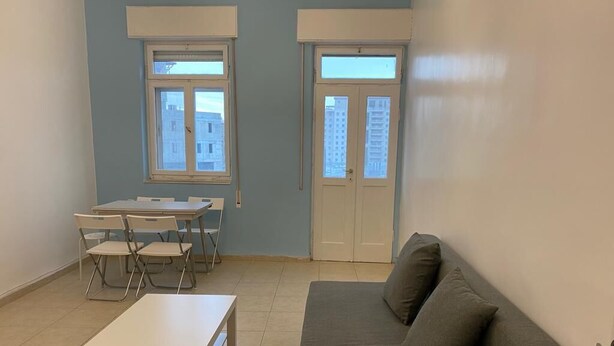 Une pièce avec un canapé gris, une petite table blanche et une table à manger avec des chaises. Il y a deux fenêtres et une porte.