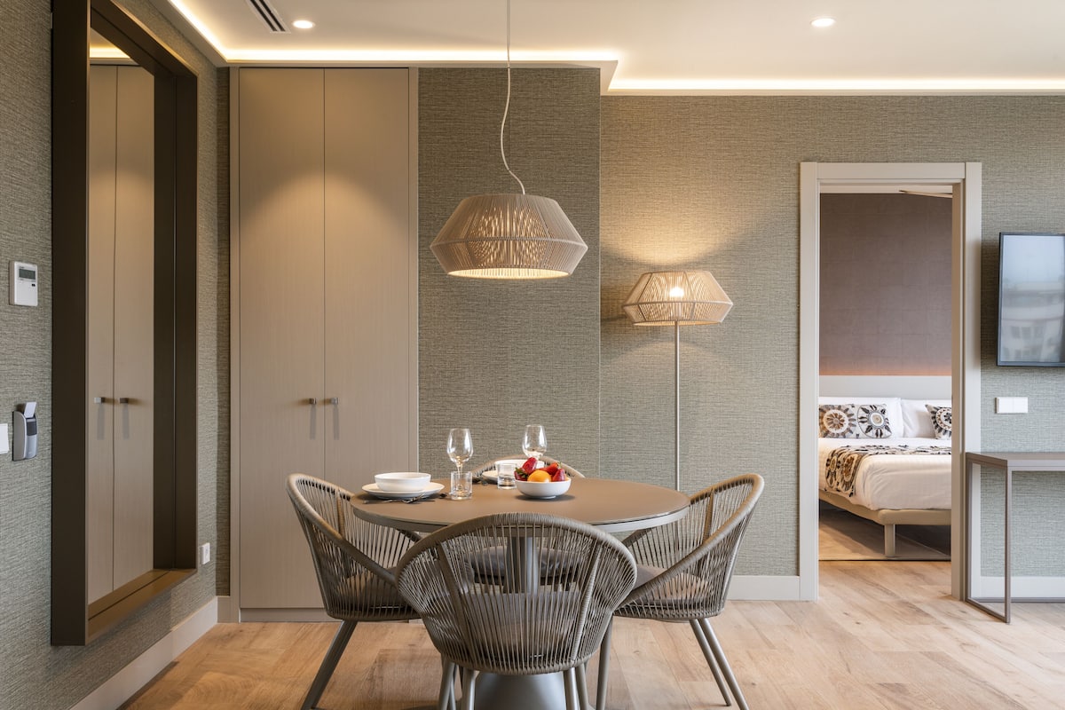 A modern dining area with a round table, wicker chairs, and a bowl of fruit.
