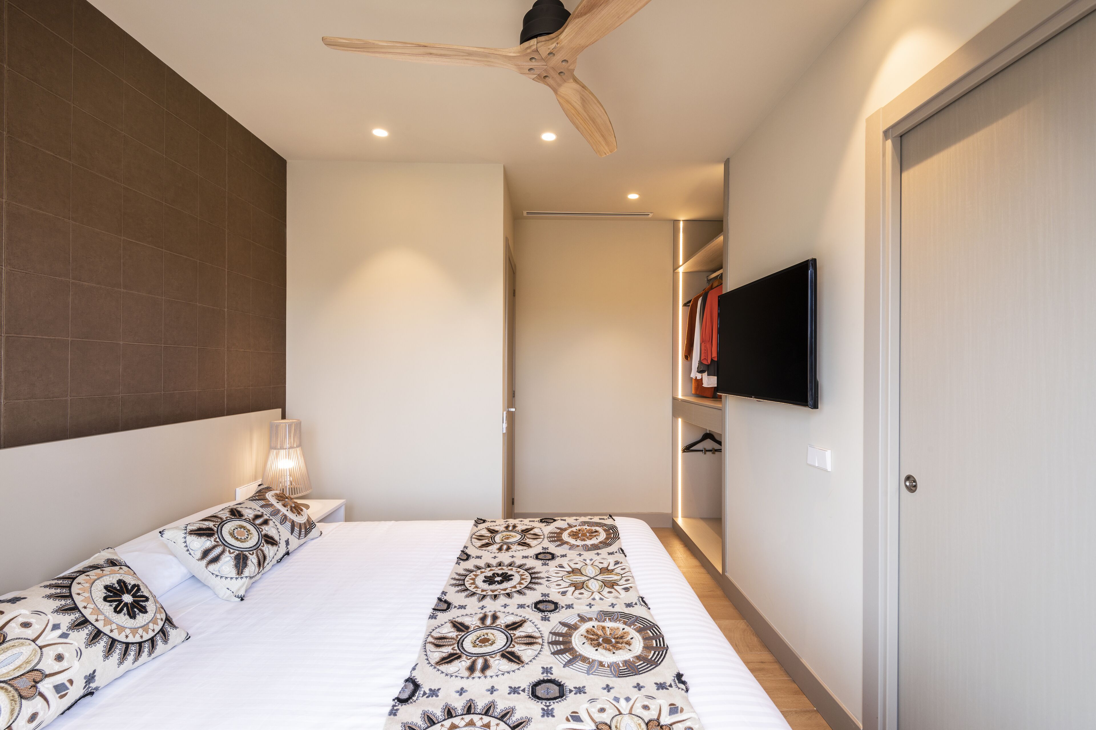 A bedroom with a bed, patterned bedspread, bedside lamps, a ceiling fan, a TV mounted on the wall, and a wardrobe.