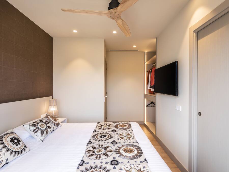 Un dormitorio con cama, colcha estampada, lámparas de noche, ventilador de techo, televisor montado en la pared y armario.
