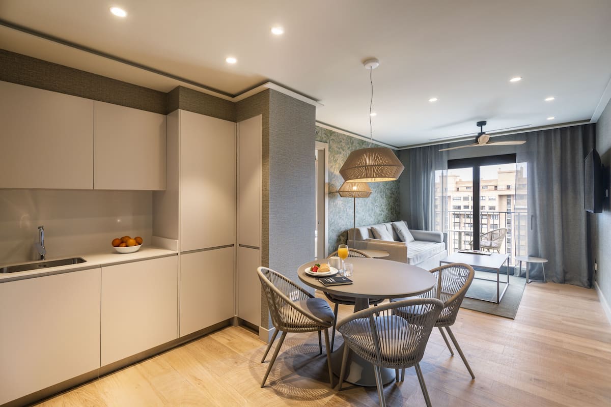 A modern kitchen with white cabinets, a dining area with a round table and wicker chairs, and a living area with a sofa and a TV.