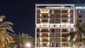 Un hôtel à plusieurs étages au design moderne, doté de balcons à chaque niveau et d’une enseigne bien visible indiquant « HÔTEL V ».