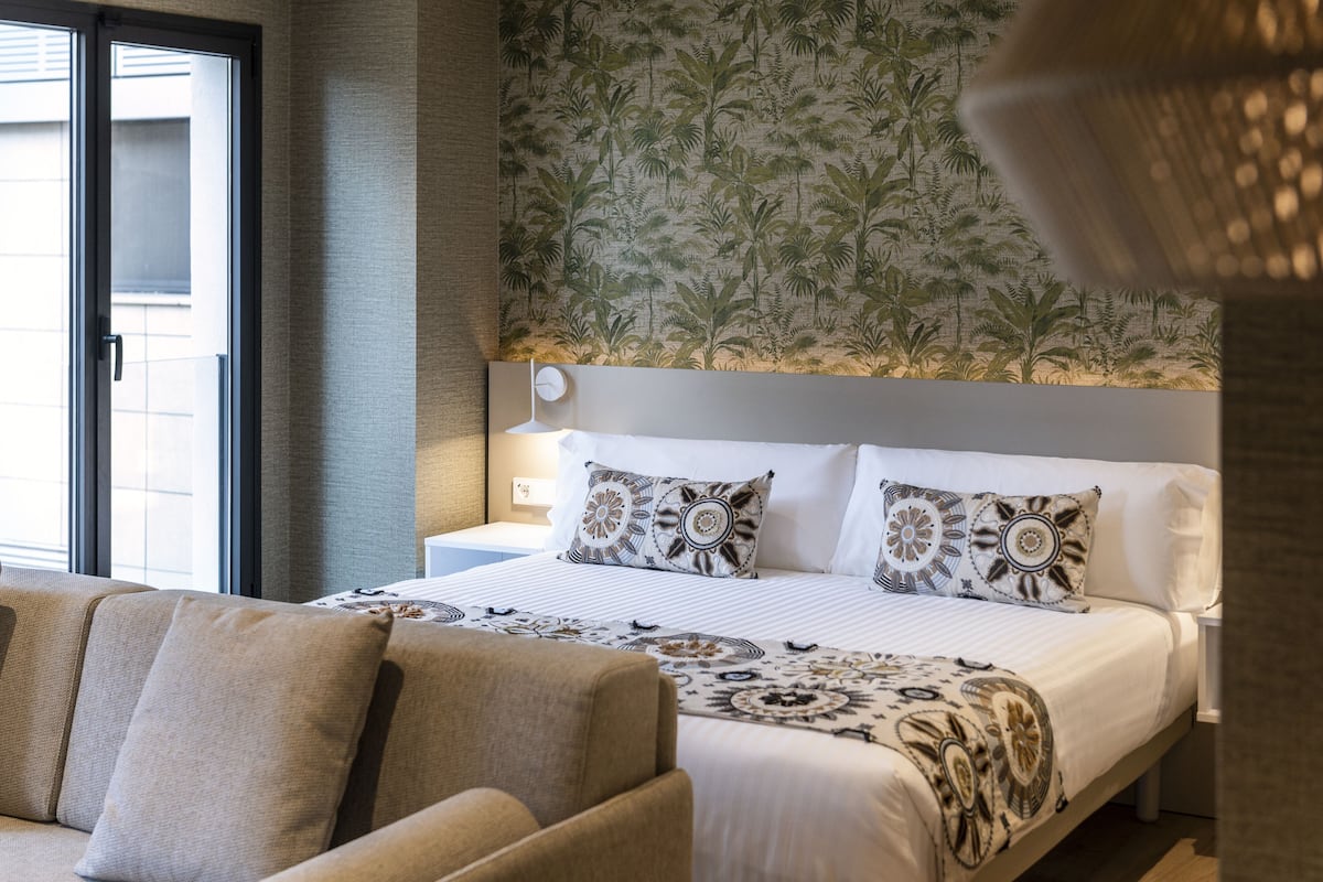 A hotel room with a bed, a sofa, a patterned wall, and a window.