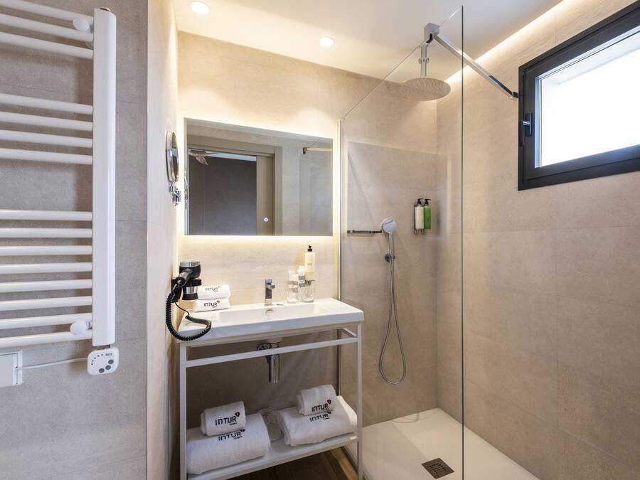 Un baño moderno con ducha envuelta en vidrio, un lavamanos blanco y un perchero con toallas dobladas con cuidado.