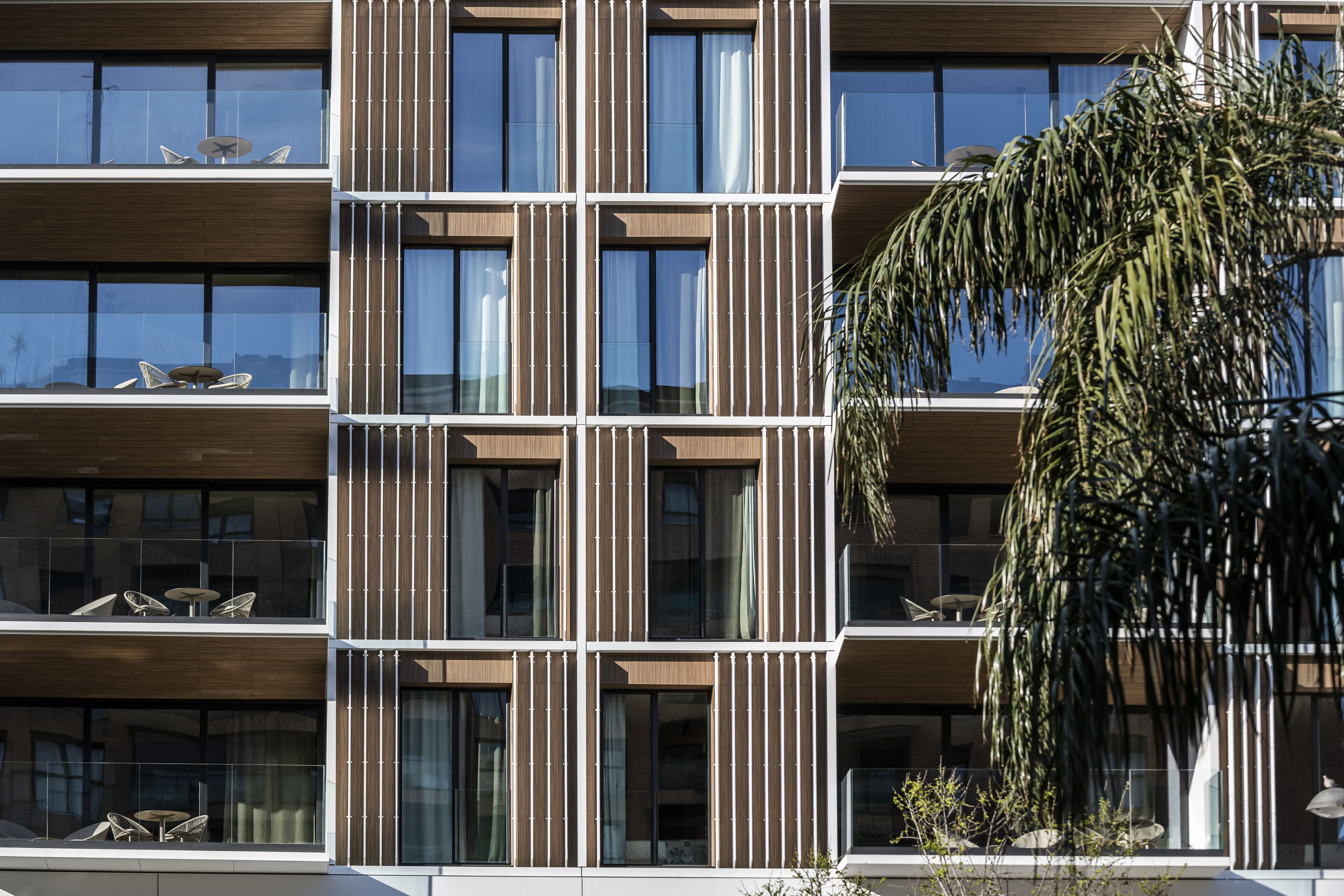A multi-story building with a modern design, featuring balconies with outdoor furniture and large windows.