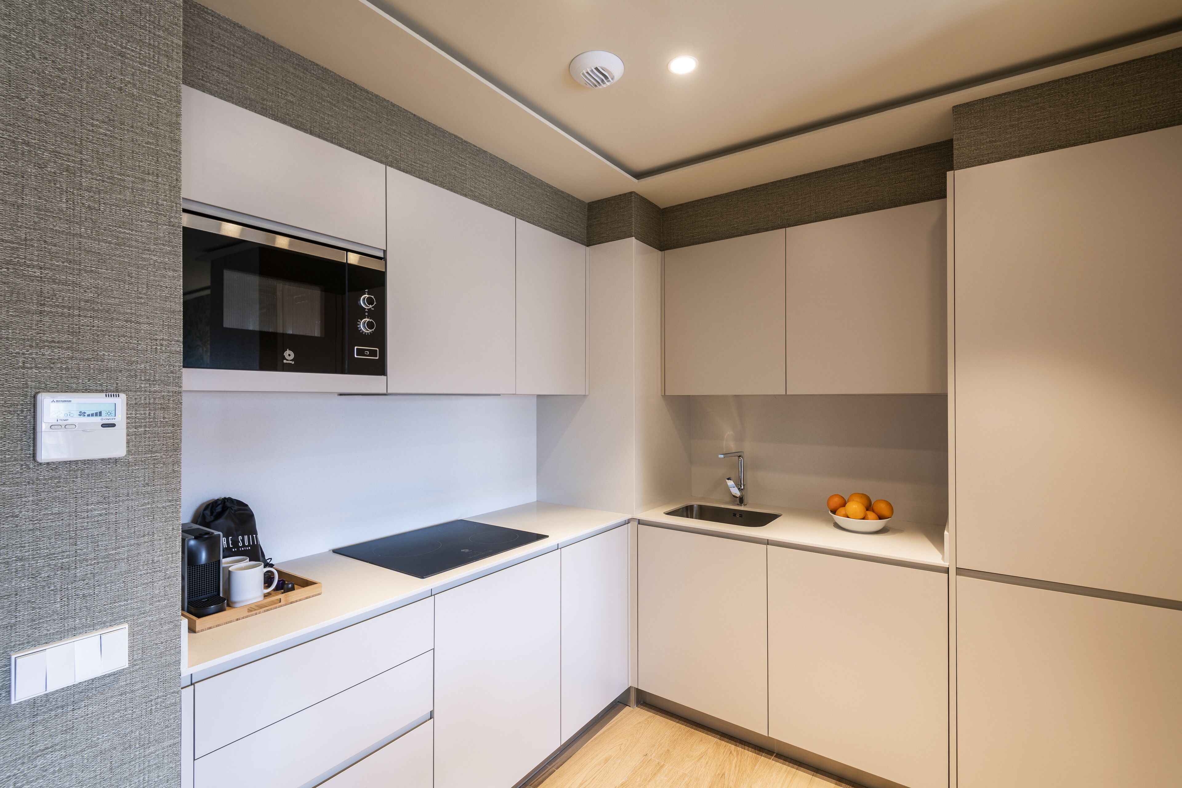 A modern kitchen with white cabinetry, a black microwave, a coffee machine, and a bowl of oranges on the counter.