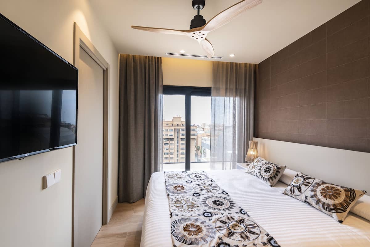 A modern hotel room with a large bed, a flat-screen TV, a ceiling fan, and a view of the city.