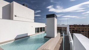 Une piscine sur le toit, dotée d’une terrasse en bois, entourée de bâtiments modernes et surplombant un ciel dégagé.
