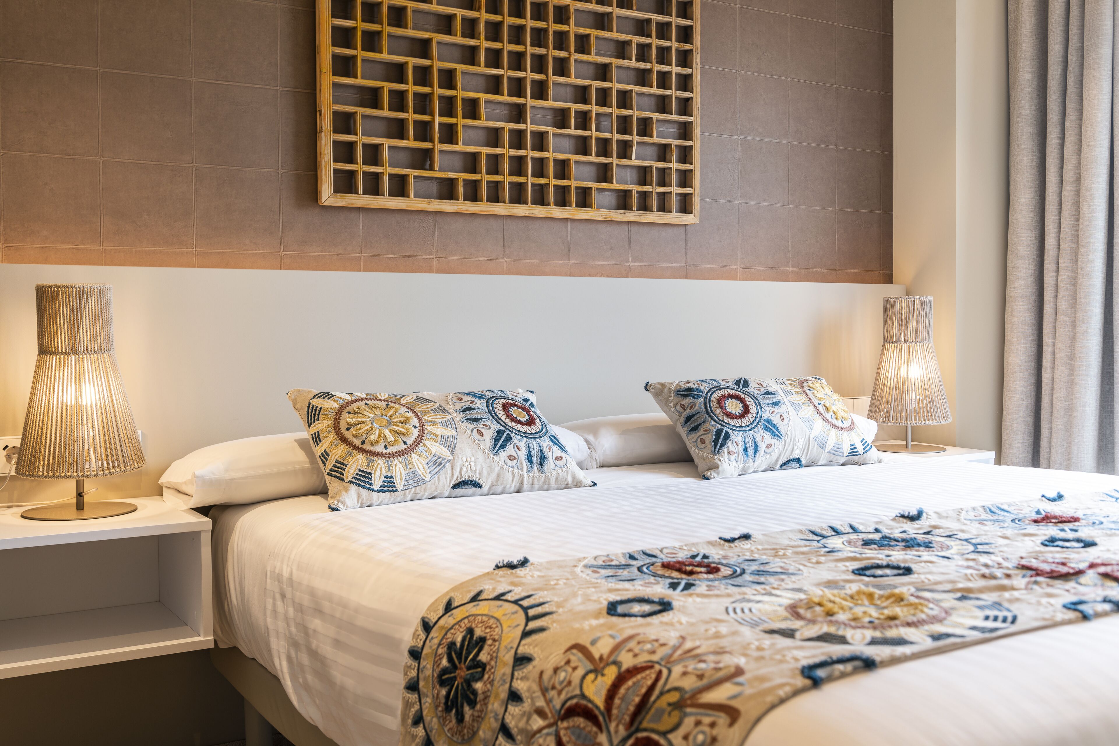 A hotel room with a large bed, two bedside lamps, a decorative wall panel, and a window with curtains.