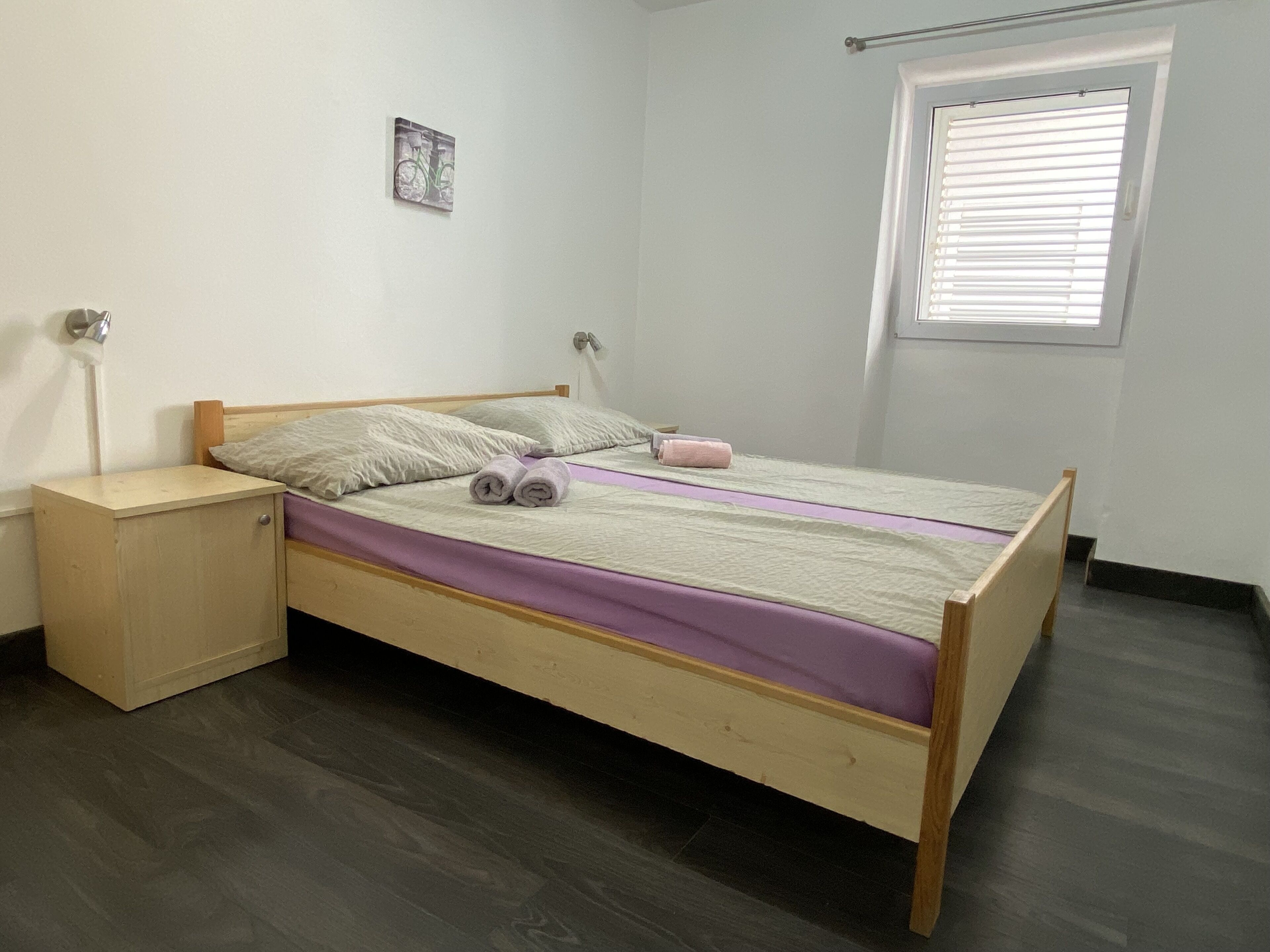 A single bed with a wooden frame, a bedside table, a small cabinet, a window with blinds, and a wall-mounted picture.