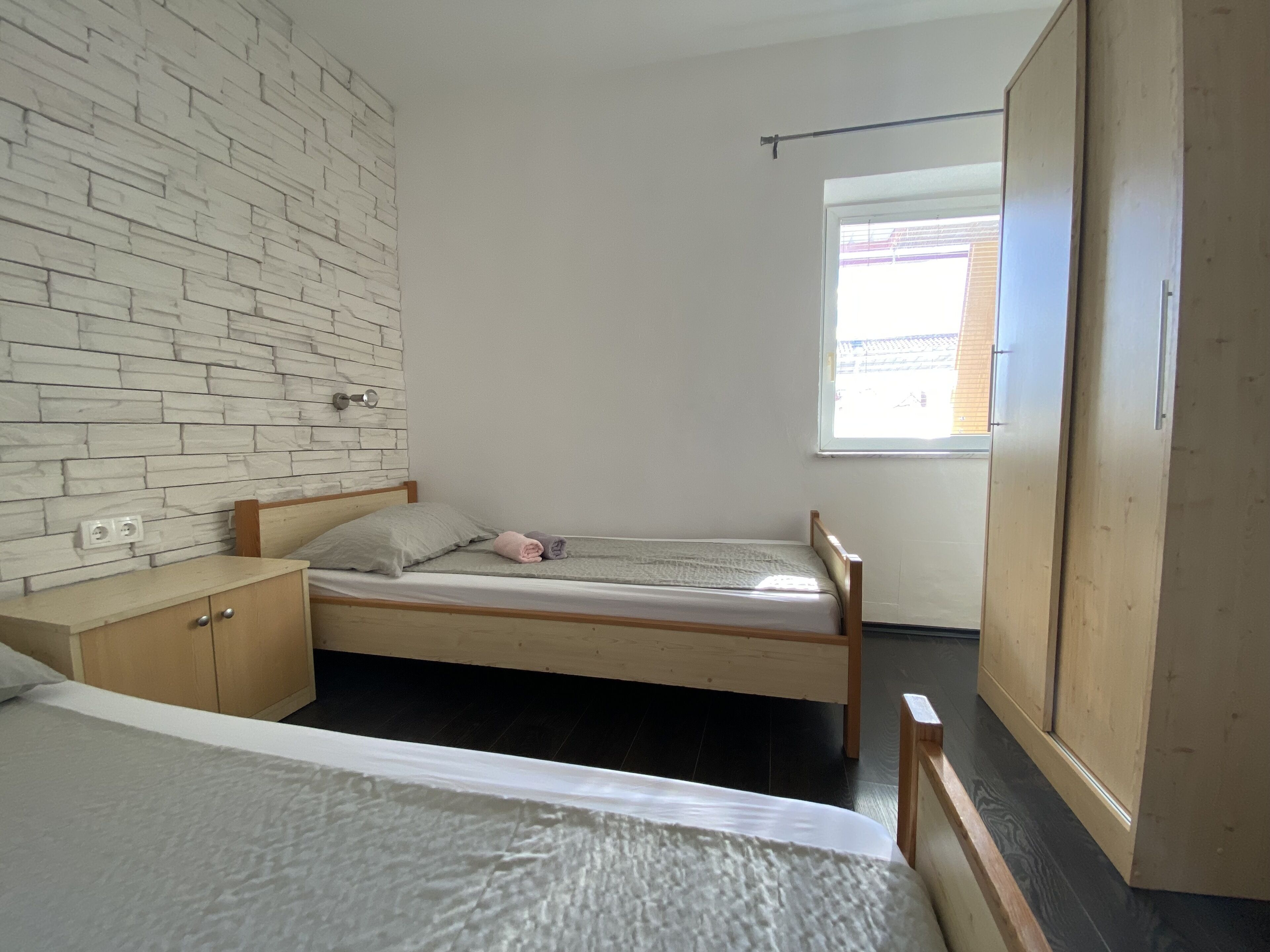 Un dormitorio con cama, armario de madera, un mueble pequeño, una ventana y pared con diseño de ladrillo.